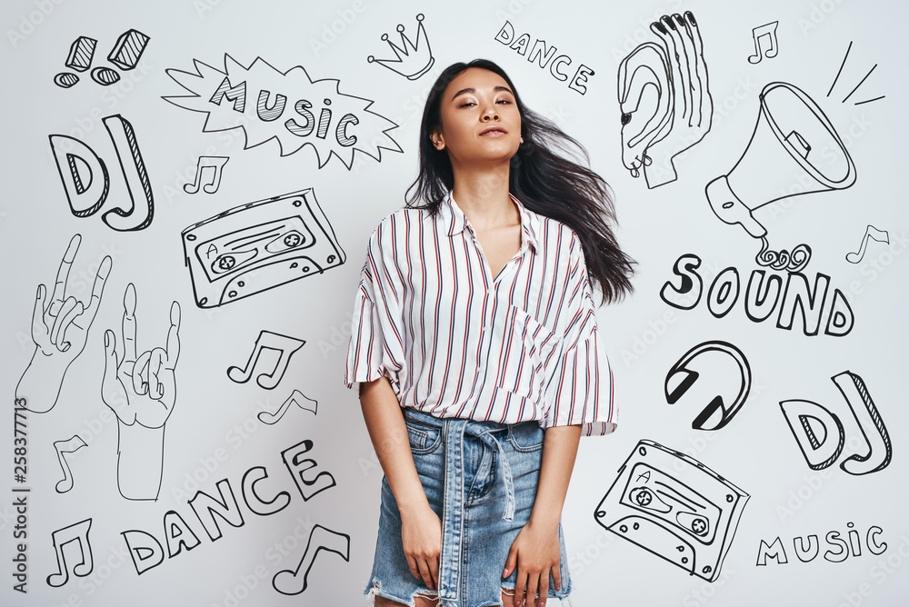 Fototapeta premium Relaxing with my favourite music...Young charming asian woman in stylish wear is looking at camera while standing against grey background with music theme doodles