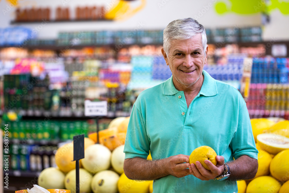 Fototapeta premium Handsome man posing, looking at camera and holding melon in hand. Bearded customer smiling. Section with fresh citruses on background