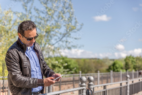 A man in leather jacket with black glasses and blue shirt reviews some issues on his mobile phone