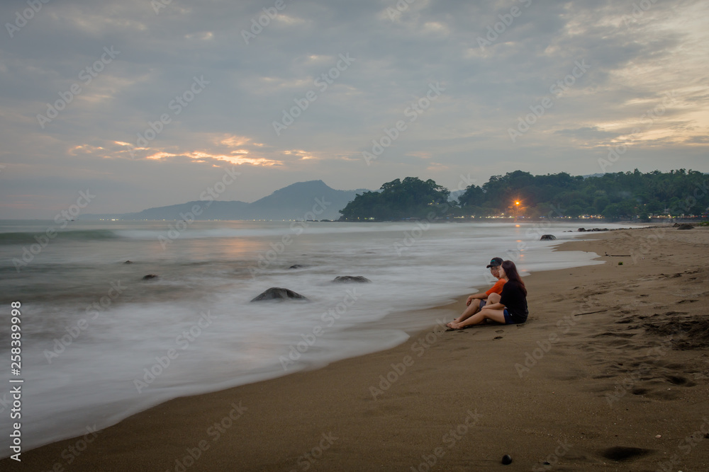 Happy couple enjoying their moments on Karang Hawu Beach, West Java, Indonesia with an orange sunset sky and blurry smooth waves