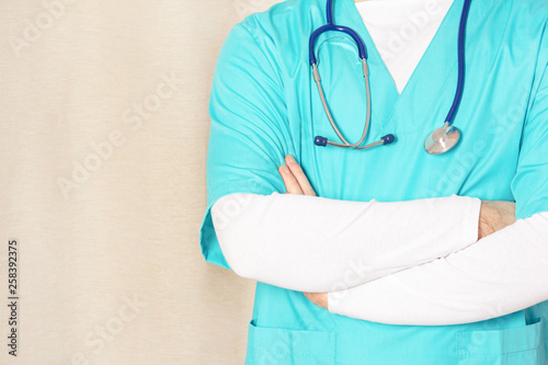 A man in a bright turquoise medical uniform folded his arms across his chest, next to a copy space. Cropped photo of paramedic, closeup. Bright photo with space for text.