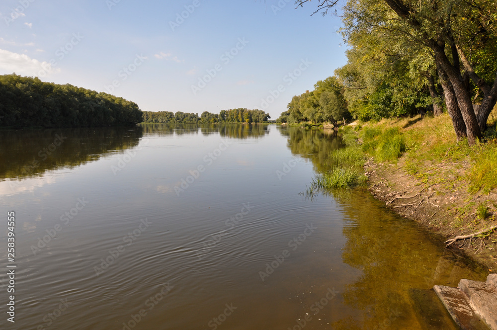 Wide yellow river is slow flowing. Early autumn. Sunny weather and brightgreen trees on the banks fron the both sides Wide yellow river is slow flowing. Early autumn. Sunny weather and brightgreen trees on the banks fron the both sides