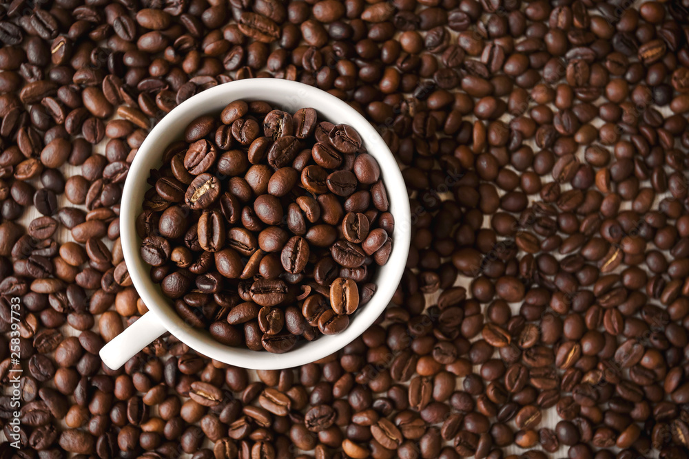 white cup with roasted coffee beans stands on the background of a large number of coffee beans white cup with roasted coffee beans stands on the background of a large number of coffee beans