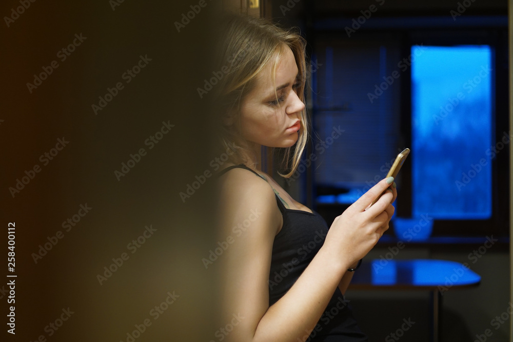 Indoor shot of European young woman holds modern phone, dressed in casual clothes, uses internet for networking and communication, stands near wall Indoor shot of European young woman holds modern phone, dressed in casual clothes, uses internet for networking and communication, stands near wall
