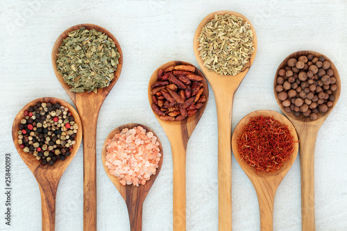 Dried herb & spice food seasoning in olive wood spoons on rustic wood. Left to right peppercorns, herbs de provence, salt, chilli peppers, fennel, saffron & allspice. 