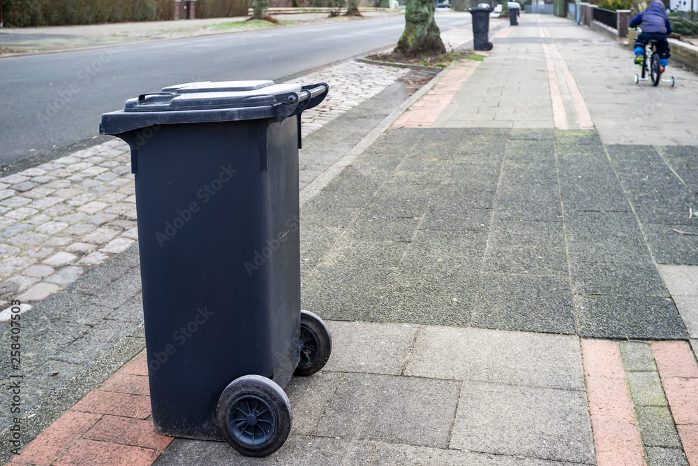 A plastic dumpster stands on the side of the road waiting for a garbage truck in the countryside, in the background a child rides a bicycle on a spring day. A plastic dumpster stands on the side of the road waiting for a garbage truck in the countryside, in the background a child rides a bicycle on a spring day.