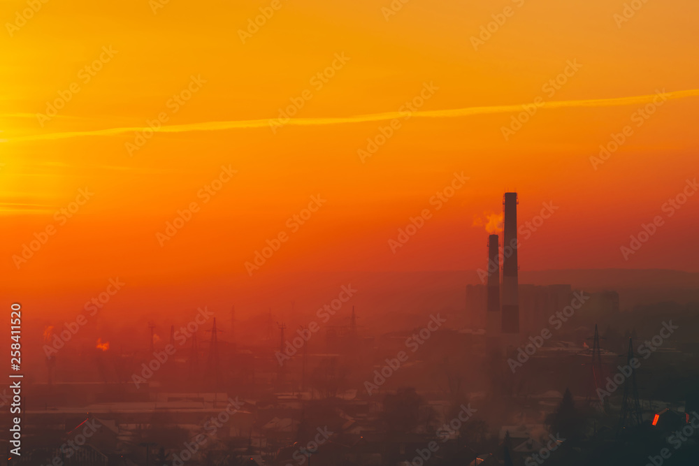 Smog among silhouettes of buildings on sunrise. Smokestack in dawn sky. Environmental pollution on sunset. Harmful fumes from stack above city. Mist urban background with warm orange yellow sky.