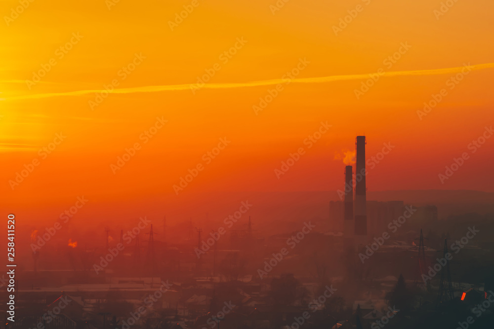 Smog among silhouettes of buildings on sunrise. Smokestack in dawn sky. Environmental pollution on sunset. Harmful fumes from stack above city. Mist urban background with warm orange yellow sky. Smog among silhouettes of buildings on sunrise. Smokestack in dawn sky. Environmental pollution on sunset. Harmful fumes from stack above city. Mist urban background with warm orange yellow sky.