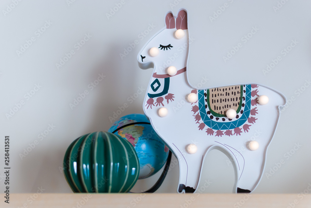 A trendy llama kids night lamp standing on a bedside table in dawn or  sunrise light with ceramic cactus and a plastic globe Stock Photo | Adobe  Stock