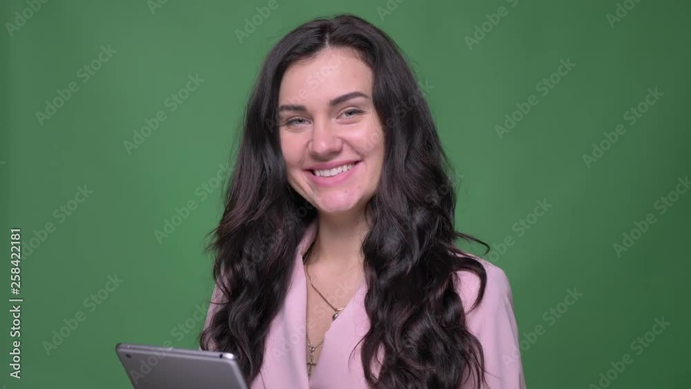 Portrait of young brunette female vloger in pink jacket working with tablet turns to camera and smiles on green background. Portrait of young brunette female vloger in pink jacket working with tablet turns to camera and smiles on green background.