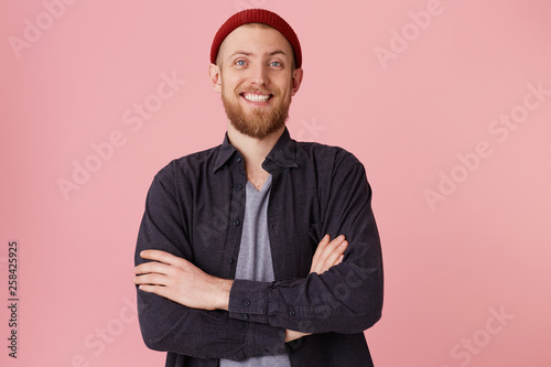 Isolated shot of joyful bearded young cute man laughs joyfully, wears red hat, with folded arms,looking at the camera over pink background. Happiness and positive emotions concept. photo