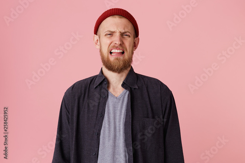Ugh, how disgusting! Displeased young bearded male, with red hat, says fie and has dissatisfied facial expression as sees something abominable or detestable, looking at camera over pink background.