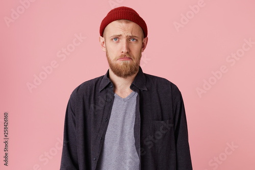 Close up of a sad bearded man about to cry, in blak shirt and t-shirt, with red hat, looking at the camera and frowning over pink background. photo