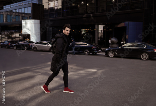 Young man crossing the road of city street. Wearing in black jacket and trousers, red sneakers, backpack, headphones. Fashion style