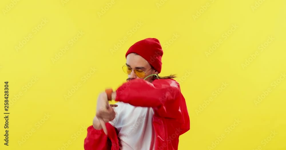Caucasian young handsome hipster man in the red hoodie, hat and sunglasses having fun and showing off in front of the camera on the yellow back wall. Portrait shot. Caucasian young handsome hipster man in the red hoodie, hat and sunglasses having fun and showing off in front of the camera on the yellow back wall. Portrait shot.