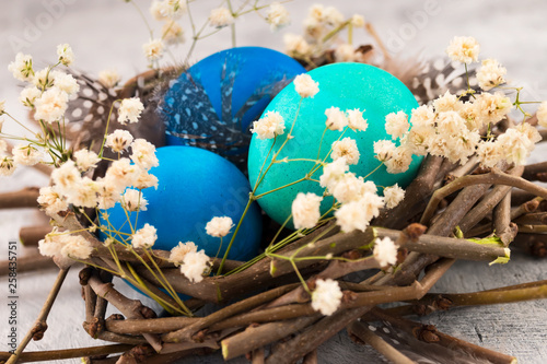 Green and blue Easter eggs in a bird's nest with white flowers and feathers. Decoration for Easter on a light background.