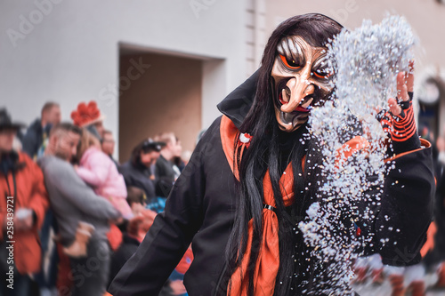 Terrible witch in black and orange robe throws white confetti. Street Carnival in Southern Germany - Black Forest.