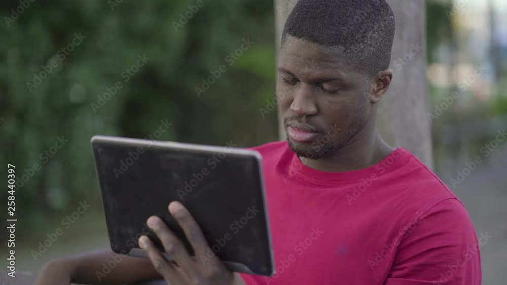 Medium shot of young Afro-American short-cut muscular man in rose T-shirt sitting on bench in park, watching football match on tablet, feeling disappointed, massaging head. Lifestyle, sport concept Medium shot of young Afro-American short-cut muscular man in rose T-shirt sitting on bench in park, watching football match on tablet, feeling disappointed, massaging head. Lifestyle, sport concept