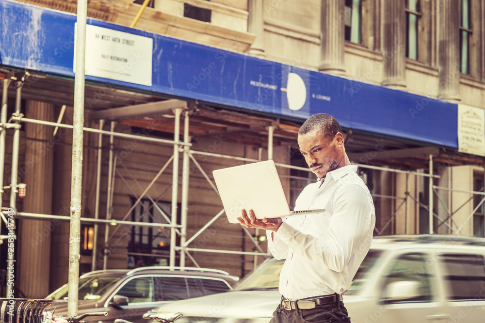 Young African American businessman traveling, working in New York City, wearing white shirt, hands holding laptop computer, walking on street, looking down, reading. Cars, buildings on background..