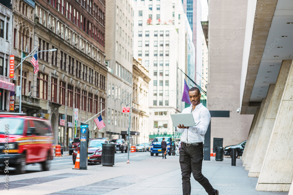 Obraz premium Young African American businessman traveling, working in New York, wearing white shirt, black pants, holding laptop computer, walking on street, looking down, reading. Cars, buildings on background.