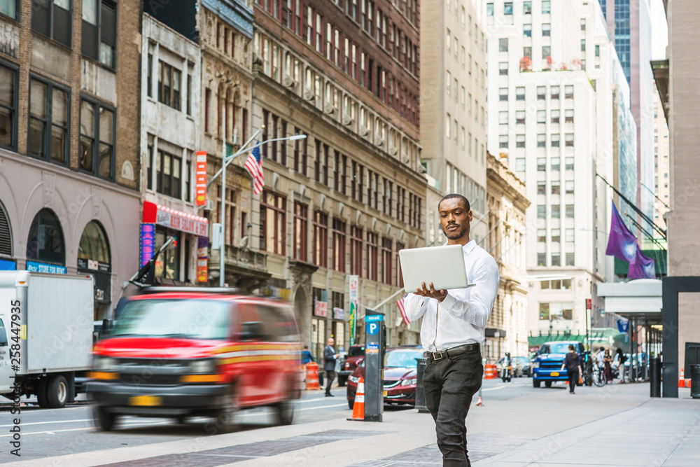 Young African American businessman traveling, working in New York, wearing white shirt, black pants, holding laptop computer, walking on street, looking down, reading. Cars, buildings on background.
