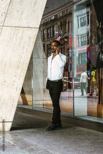 Young African American businessman working in New York, wearing white shirt, black pants, leather shoes, carrying laptop computer, standing outside office building, talking on cell phone, thinking..