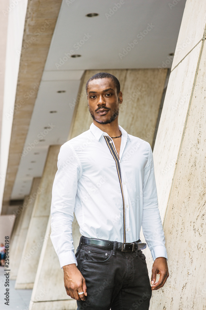 Portrait of Young African American Man in New York City. Young black guy  with beard, wearing white long sleeve shirt, black pants, standing outside  office building, confidently looking forward.. Stock Photo