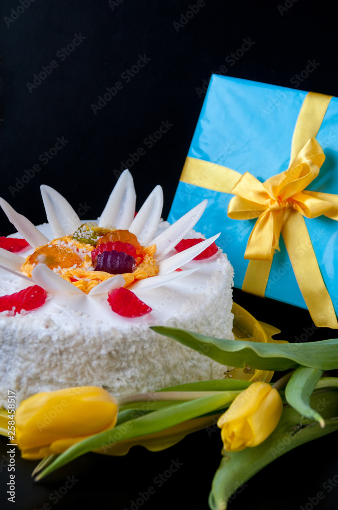 White cake, decorated with petals and jelly, yellow tulips and a gift on a black background
