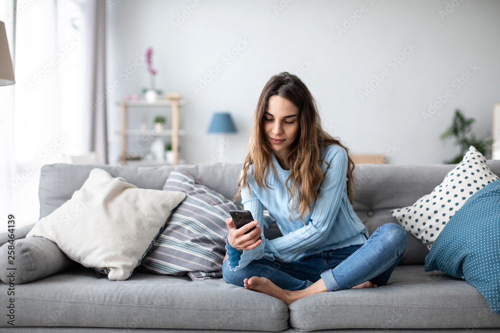 Attractive smiling woman using smart phone while sitting on the sofa at home. - obrazy, fototapety, plakaty 