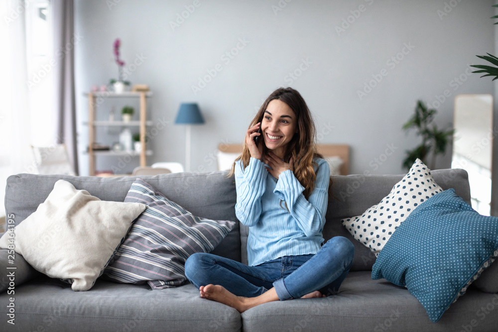 Smiling woman talking on the phone at home on the sofa. - obrazy, fototapety, plakaty 