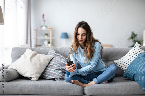 Attractive smiling woman using smart phone while sitting on the sofa at home.