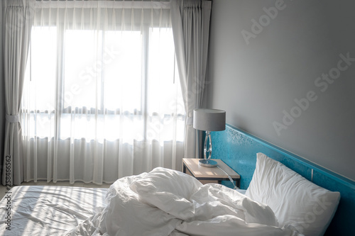 Selective empty unmade messy bed in the morning. White bedroom, bed, pillow and curtain with blue wallpaper in the morning, sunlight shining through the window shade after wake up. photo