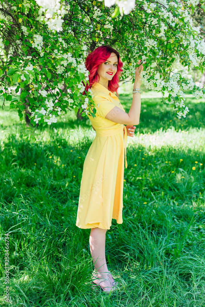 Beautiful and young woman with bright red hair and ref lips standing next to a blooming apple tree in a yellow dress. Beautiful and young woman with bright red hair and ref lips standing next to a blooming apple tree in a yellow dress.