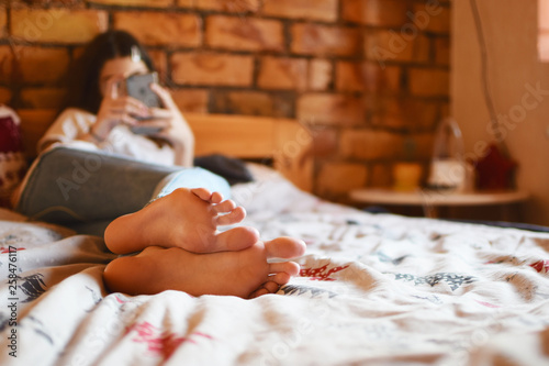 Teenage girl blogger using gadget smartphone or mobile phone texting message using wifi internet while lying on bed in her room with red brick wall, shallow depth of field with focus at bare feet.