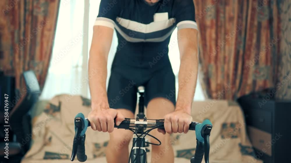 Man Trains On A Bicycle Simulator.Man Training Cycling Bicycle Indoor.Man In A Black Shirt On Exercise Bike At Home.Guy Exercises On The Bike At Home.Man's Working Out On The Exercise Bike. 