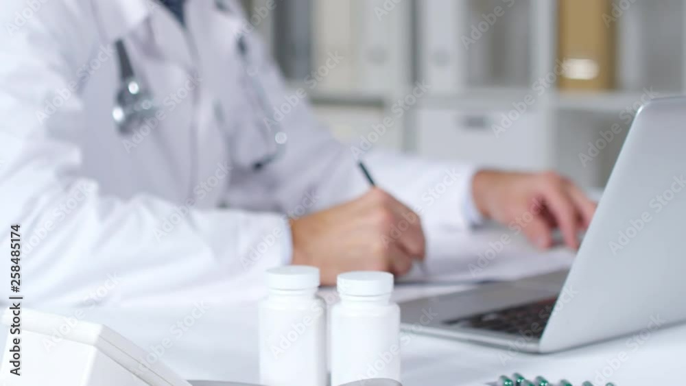 Tilt up shot of busy male medical specialist in white coat sitting at his desk and filling form on clipboard Tilt up shot of busy male medical specialist in white coat sitting at his desk and filling form on clipboard