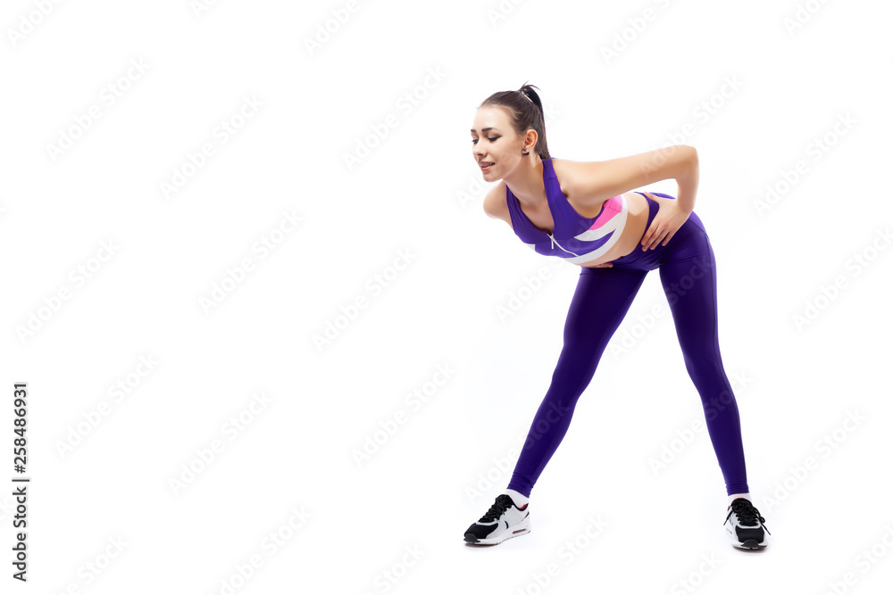 A dark-haired woman coach in a sporty purple  short top and gym leggings shows the correct technique of tilting forward for inflating the buttocks on a  white isolated background in studio