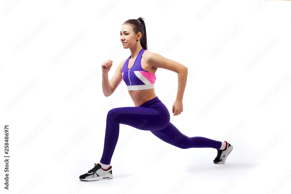 A dark-haired woman coach in a sporty purple  short top and gym leggings makes lunges  by the feet forward, hands are held out to the side   on a  white isolated background in studio