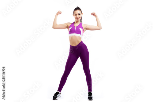 A dark-haired woman coach in a sporty pink short top and gym leggings shows biceps and smiles  on a  white isolated background in studio © Виталий Сова