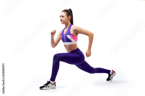 A dark-haired woman coach in a sporty purple  short top and gym leggings makes lunges  by the feet forward, hands are held out to the side   on a  white isolated background in studio © Виталий Сова