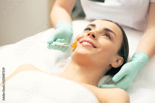Young lady smiling while cosmetologist using peeling brush