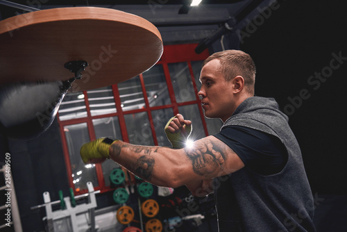 Stay strong to win. Side view of professional young boxer with green hands bandages hitting punching speed bag while training his boxing skills in boxing gym
