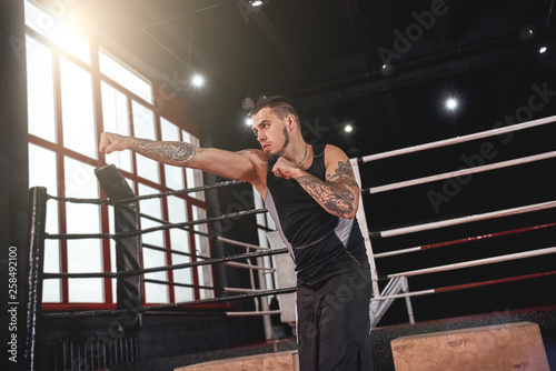 Training hard. Muscular athlete in sports clothing throwing right cross in boxing gym