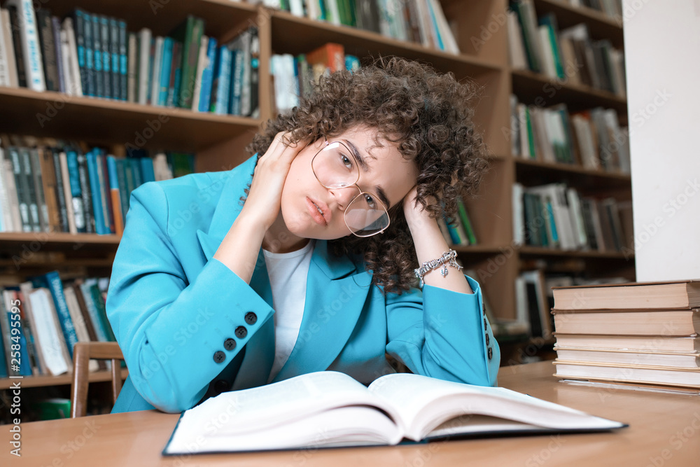 Young beautiful curly girl in glasses and blue suit sitting with books in the library. Student Study Young beautiful curly girl in glasses and blue suit sitting with books in the library. Student Study