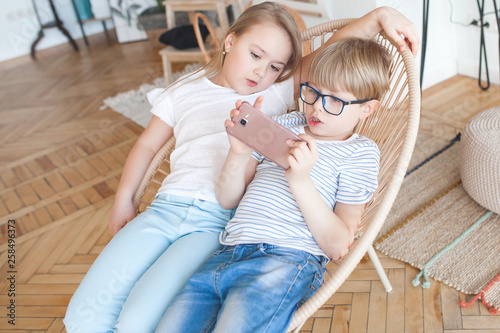 Two kids playing smartphone. Boy and girl at home watching at the cell phone. Digital children indoor. photo