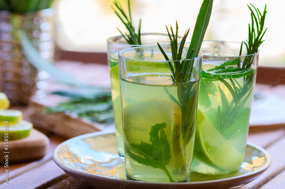 Fototapeta premium Three glasses with atural lemonade made from lemon rosemary and mint in glasses on wooden table at the terrace. Cold refreshing beverage for hot summer day.