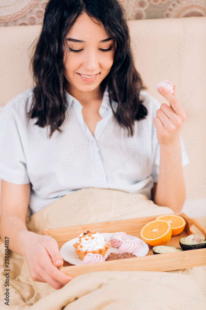 Beautiful awesome dark haired woman having tasty breakfast in bed at her cozy bedroom. Side view photo of young girl in blue striped pajamas eating sweets. Beautiful awesome dark haired woman having tasty breakfast in bed at her cozy bedroom. Side view photo of young girl in blue striped pajamas eating sweets.