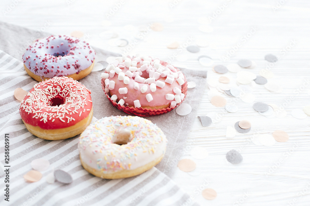 Delicious colorful donuts with sprinkles and marshmallows on stylish white table with confetti, copy space. Party concept. No diet. Candy bar at wedding reception. Purple, pink donuts