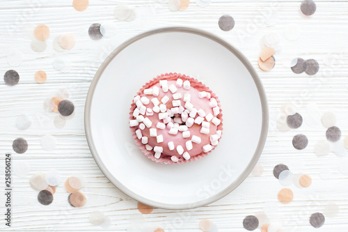 Delicious colorful donut with sprinkles and marshmallows on stylish plate on white table with confetti, flat lay. Party concept. No diet. Candy bar at wedding reception. Pink donut