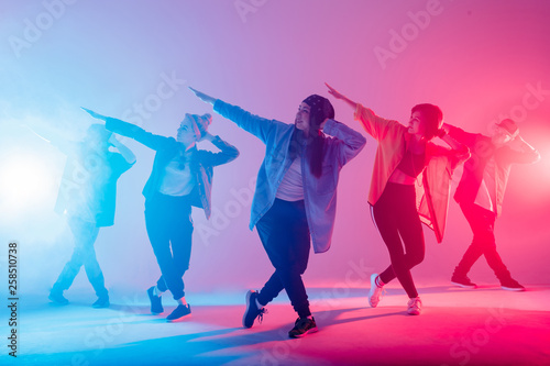 Fototapeta Naklejka Na Ścianę i Meble -  Young modern dancing group of six adult young people practice dancing on colorful background. Fashionably dressed youngsters moving over blurred disco club color lights